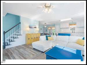 Bright, modern living room with white sectional sofa, blue accents, hardwood floors, and an adjacent kitchen. features include a ceiling fan and a staircase on the left.