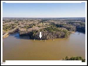 Aerial view of a lake with a wooded shoreline. A white arrow points to a specific dock along the water's edge at 490 Flanagan Road. Several other docks are visible along the shoreline, surrounded by dense forest and sparse buildings in Henderson. The water is calm under a clear sky.