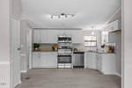A modern kitchen with white cabinets, stainless steel appliances, and grey flooring. there's a wooden dining table on the right and bright natural light coming through a window above the sink.