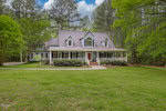 A large two-story house with a gray roof, lavender siding, and red door, featuring a wide, covered front porch and surrounded by dense green trees. the house has a well-manicured lawn in a forested area.