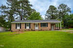 Located at 434 Lightwood Lane in Henderson, NC, this charming brick single-story house features white trim, four front windows, and a small front porch with a white door. A carport is attached to the right. The house is nestled among tall pine trees and surrounded by a grassy lawn under a partly cloudy sky.