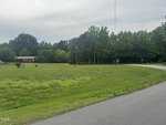 A green lawn with a few scattered trees and shrubs extends to a house in the background surrounded by more trees. In the foreground, a paved road curves to the right, with power lines and a stop sign along the roadside. The sky is cloudy and overcast.