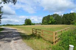 A scenic view of a rural landscape featuring a gravel road running alongside a grassy, fenced-in field. The field is bordered by a line of tall, green trees under a partly cloudy blue sky. A small building is visible in the distance, surrounded by greenery.
