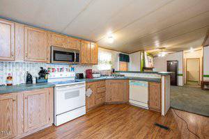 A kitchen with wooden cabinets, green countertops, and a white stove with a microwave above it. There is a dishwasher to the right and various kitchen appliances on the countertops. The floor is hardwood, and there are two small windows with curtains.