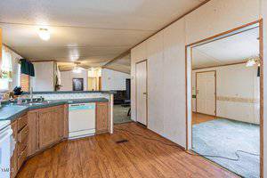 A mobile home interior with a wood-floored kitchen featuring wooden cabinets, a dishwasher, and a stove. The hallway leads to a living area with a sofa and carpeted flooring. To the right, an open doorway reveals another carpeted room.