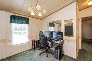 A home office setup features a desk with a computer, dual monitors, a gaming chair, and various peripherals. A large window with green curtains allows natural light to enter. The room has beige walls, a green carpet, and a chandelier hanging from the ceiling.