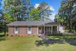 A brick single-story house with white trim and a front porch at 220 Old Watkins Road, Henderson NC. A curved sidewalk leads to the entrance. The lawn is well-maintained, and tall trees surround the property. An American flag is displayed next to the porch under a partly cloudy sky.