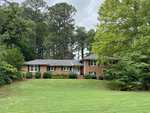 A single-story brick house with a wide front lawn, surrounded by tall trees and shrubs. The house features a central front entrance with a small staircase leading up to it, and has a well-maintained green lawn in the foreground.