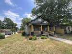 A single-story gray house with a dark gray roof sits on a corner lot at 701 S Williams Street, Henderson NC. It has a covered front porch with wooden pillars and a ramp leading up to the front door. The yard is mostly grass with a few small bushes, and large trees provide shade in the background.