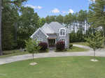 A two-story stone house at 55 Clare Drive, Henderson NC, with a light gray shingled roof is surrounded by tall trees. The house features large windows and a wooden balcony above the front porch. A circular driveway with small trees and neatly trimmed grass leads to the front entrance.