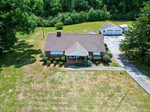 Aerial view of a single-story brick house with a gabled roof, manicured lawn, and shrubbery along the front. The property includes a detached garage and is surrounded by trees and greenery. A concrete driveway leads up to the garage from the main road.