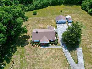 An aerial view of a property with a single-story brick house featuring a pitched roof. Adjacent to the house is a detached garage and a smaller shed. The house is surrounded by a well-kept lawn with several trees and shrubs, and a paved driveway leads to the garage.