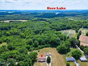 An aerial view showing a house marked by a red star amidst a large expanse of green trees and fields. Multiple other houses and clearings are visible. In the distance, Kerr Lake is labeled and can be seen under an open sky with scattered clouds.