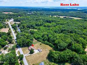 Aerial view of a rural area near Kerr Lake with dense green forests and scattered houses. A road runs diagonally across the image with a red marker indicating a property. Kerr Lake is visible in the distance under a clear, blue sky.