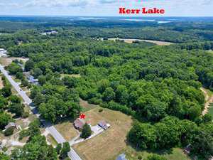 Aerial view of a rural area with a forest and scattered houses. A labeled red star marks a house in the lower left. The background shows extensive green woodlands and a large waterbody labeled "Kerr Lake." Several roads and fields are visible around the area.