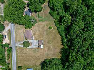 An aerial view of a property featuring a house with a brown roof, a driveway leading to the road, and a large grassy yard bordered by dense green trees. The house is surrounded by greenery, and a smaller structure is visible near the house.