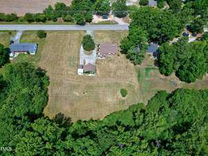 An aerial view of a rural property shows a large, clear section of land with scattered trees. The property includes a single-story house, an outbuilding, and a gravel driveway. It is bordered by a road at the top and surrounded by lush green trees and other properties.
