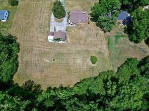 Aerial view of a rural property featuring a large lawn, a single-story house with a reddish-brown roof, a smaller outbuilding, and a curved driveway. Trees border the property, with a dense forested area at the bottom of the image.