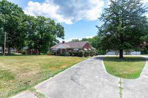 A single-story brick house with a neatly trimmed lawn is seen from a driveway that curves towards the front entrance. The house is surrounded by large trees, and the sky above is partly cloudy. A white fence is visible in the background on the right side.