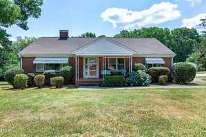 A single-story brick house with white trim surrounded by neatly trimmed shrubs and bushes. There are two large windows on either side of the front entrance, which features a small covered porch and bushes with blooming flowers. The sky is clear with a few clouds.