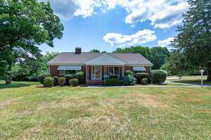 Single-story red brick house with a gabled roof, covered front porch, and awnings over the windows. Surrounded by well-manicured lawn, shrubs, and trees. Driveway is visible to the right, with a mailbox by the lawn. The sky is partly cloudy.