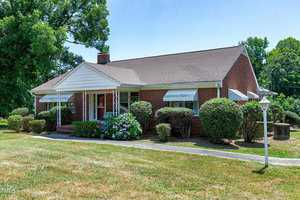 A single-story red brick house with a triangular white porch roof supported by decorative metal posts. The home is surrounded by neatly trimmed bushes and a well-maintained lawn. A white lamp post is positioned near the walkway leading to the front entrance.