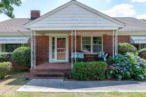 A one-story brick house with a front porch supported by decorative metal columns. The porch is furnished with a wicker loveseat, a chair, and a small table. Bushes and blooming hydrangeas surround the walkway leading to the front door. The sky is clear and sunny.