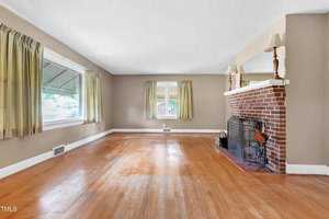 A spacious living room with hardwood floors, beige walls, and large windows with yellow curtains. There is a red brick fireplace with a black metal guard on the right, topped with a mirror and lamps. Natural light fills the room, creating a warm atmosphere.