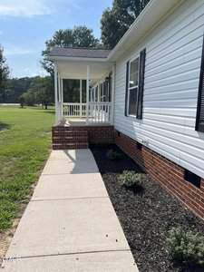 A concrete pathway leads to the entrance of a white house with black shutters. The house has a small porch with white railings and brick steps. Mulched flower beds with small shrubs line the house's foundation. The yard is grassy with trees in the background.