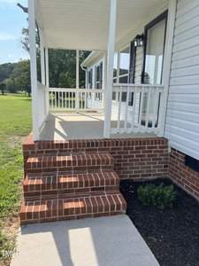 A porch with white railings and a roof extends from a white house. Red brick steps lead up to the porch, which has a light fixture near the door. The area around the steps is landscaped with small plants, and a large green lawn is visible in the background.