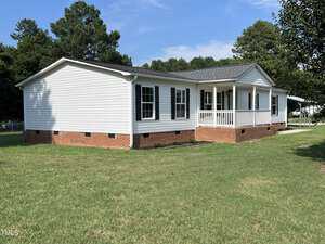 A white single-story house with black shutters, a brick foundation, and a small front porch is shown. The house is situated on a grassy lawn with trees in the background, and the sky is clear and blue.