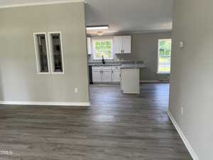 A modern, open-concept kitchen with light grey wooden flooring is shown. The kitchen features white cabinets, a granite countertop, a stainless steel sink, and large windows that let in ample natural light.