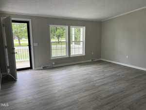 An empty living room with light gray walls, two large windows, and a door to the left that is slightly open. The floor is covered with light-colored laminate wood. Outside the windows, greenery and part of a porch railing are visible.