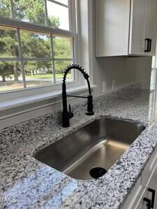 A kitchen sink with a high-arc black spring faucet set in a countertop made of speckled granite. The countertop extends along the wall below a large window, providing a view of a grassy yard with trees. White cabinets with black handles are seen on the right side.