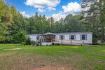 A white mobile home with a wooden ramp for accessibility is situated on a spacious green lawn. The home has a front porch with a few items and outdoor seating. It is surrounded by a thick line of trees under a partly cloudy sky.