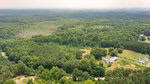 Aerial view of a lush, green rural area with dense forests, scattered clearings, and several houses. Rolling hills extend into the distance under a hazy sky, and a narrow road winds through the landscape.