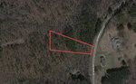 An aerial view of a wooded area in Kittrell with a triangular plot of land outlined in red. The plot is bordered by what appears to be a dirt road and two houses on the right side, along with a tree-filled landscape surrounding it. The location is near 0 Sims Bridge Road.
