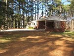 Single-story brick house in a wooded area. The building features a porch area with white railings, a large front window, and a sloping roof. The surrounding yard is covered with a mix of grass and fallen pine needles, and tall pine trees encircle the house.