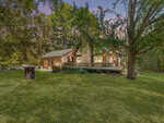 A rustic log cabin at 78 Walter Grissom Road with a stone chimney sits surrounded by tall trees. The house features a wooden deck wrapping around the front and sides. A well-kept lawn extends to the foreground, with a large tree providing partial shade. The Kittrell sky is a gradient of colors at sunset.