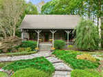 A charming wooden cabin with a front porch, surrounded by lush greenery. Stone steps and a pathway lead to the porch, which has potted plants, a porch swing, and outdoor seating. Nestled among trees at 404 General Green Road, Kittrell, the cabin is embraced by neatly landscaped gardens.