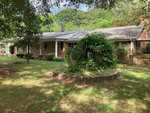 A single-story brick house at 3158 Sims Bridge Road, Kittrell, with a gray shingle roof is surrounded by lush greenery. The front yard has a circular raised garden bed with dense foliage. A wooden ramp leads to the front entrance on the right side of the house. Trees and bushes frame the scene.