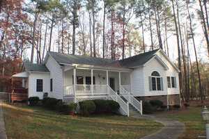 A white single-story house at 135 S S Chaucer Way, featuring a front porch and steps leading to the entrance, is situated amidst tall trees. The Kittrell residence has a gable roof and is surrounded by a well-kept lawn and bushes. The setting appears peaceful with woodland scenery in the background.