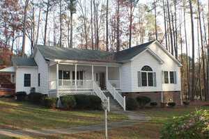 A single-story white house with a spacious front porch and a brick foundation, surrounded by tall trees and a lawn. The home features several large windows and a set of steps leading up to the front door. A curved walkway leads from the front door to the yard.