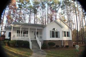 A single-story white house with a brick foundation, surrounded by tall pine trees. The house has a large porch with white railings, a cement walkway leading to a small staircase, and a swing set visible in the right background. The setting appears to be autumn.