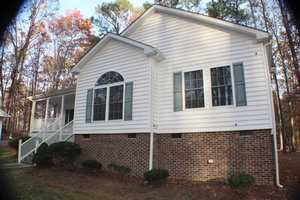 A white house with light green shutters features a combination of brick and siding exterior. The house has a large, arched window on the front and a small set of steps leading to a covered porch. It is surrounded by a grassy yard and tall trees.