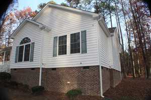 A two-story house with white siding and a brick foundation is surrounded by tall trees. The side of the house features large windows with shutters, and a downspout is visible on the right corner. The yard has trimmed shrubs and fallen leaves on the ground.