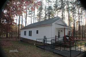 A small white building with a slanted roof, surrounded by tall trees. It has a ramp for accessibility leading to a porch with red railings. The ground is covered with dry leaves, and the scene appears to be taken during autumn.