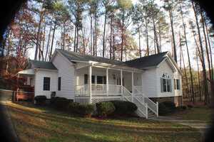 White single-story house with green shutters and a front porch with white railings and stairs. Surrounded by grass, bushes, and tall trees. A paved walkway leads up to the porch, and the house has a side addition with a small deck and lattice.