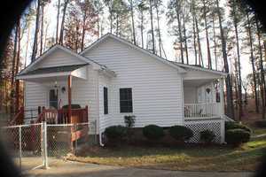 A white single-story house with a green roof is surrounded by tall trees. The home has a small front porch with railings, a covered side patio, and a chain-link fence to the left. Shrubs are neatly planted along the foundation. The setting appears peaceful.