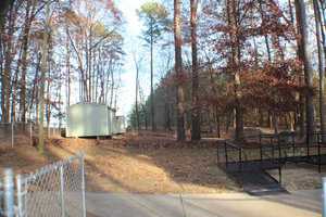 A small, light-gray shed is nestled among tall trees in a wooded area. A black ramp leads to the shed, and autumn leaves cover the ground. In the foreground, there is a chain-link fence and a concrete pathway. The sky is clear, indicating a sunny day.